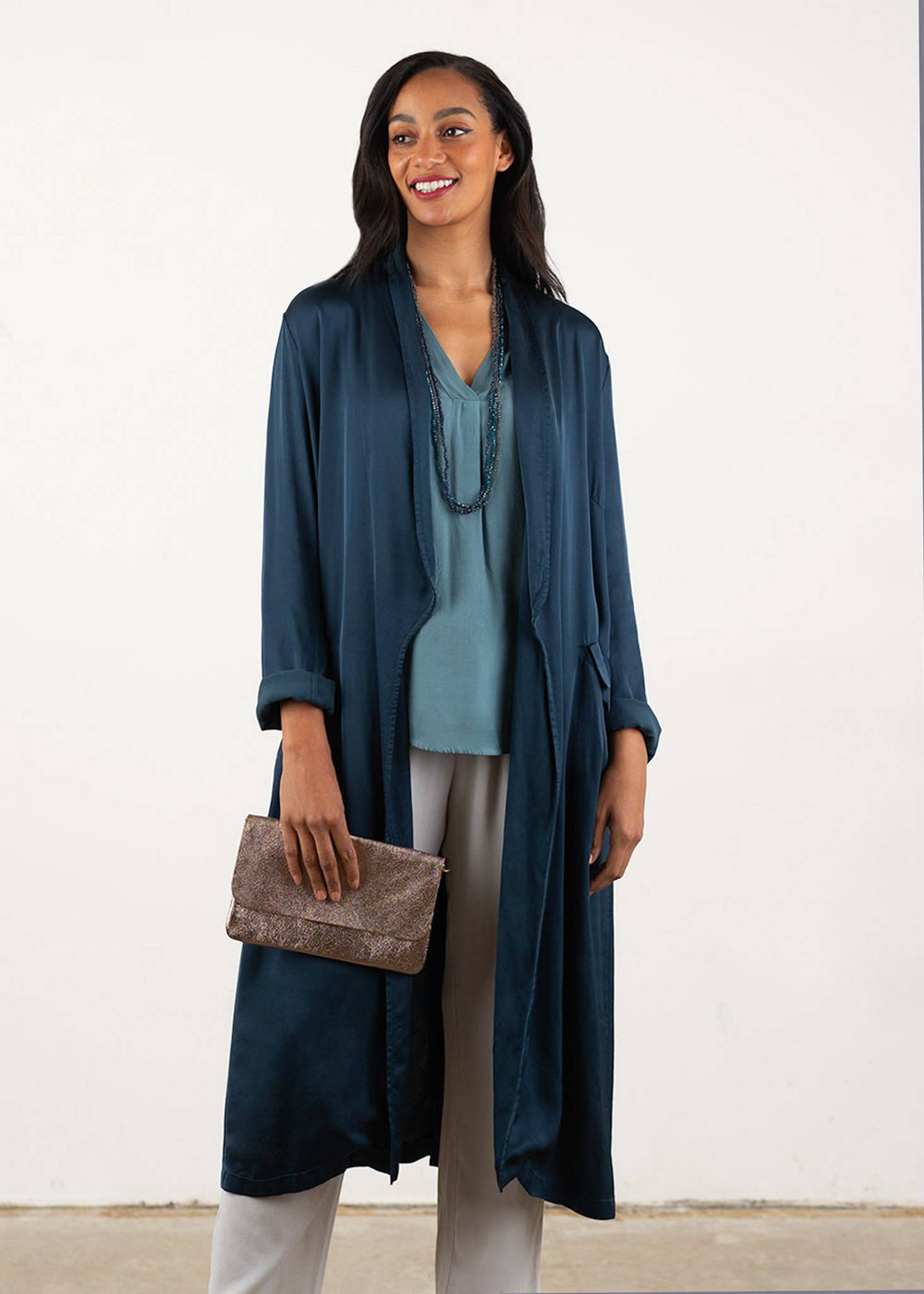 A model wearing a longline teal satin jacket over a blue grey sleeveless top, crystal necklaces, oatmeal trousers and a metallic champagne coloured clutch  bag