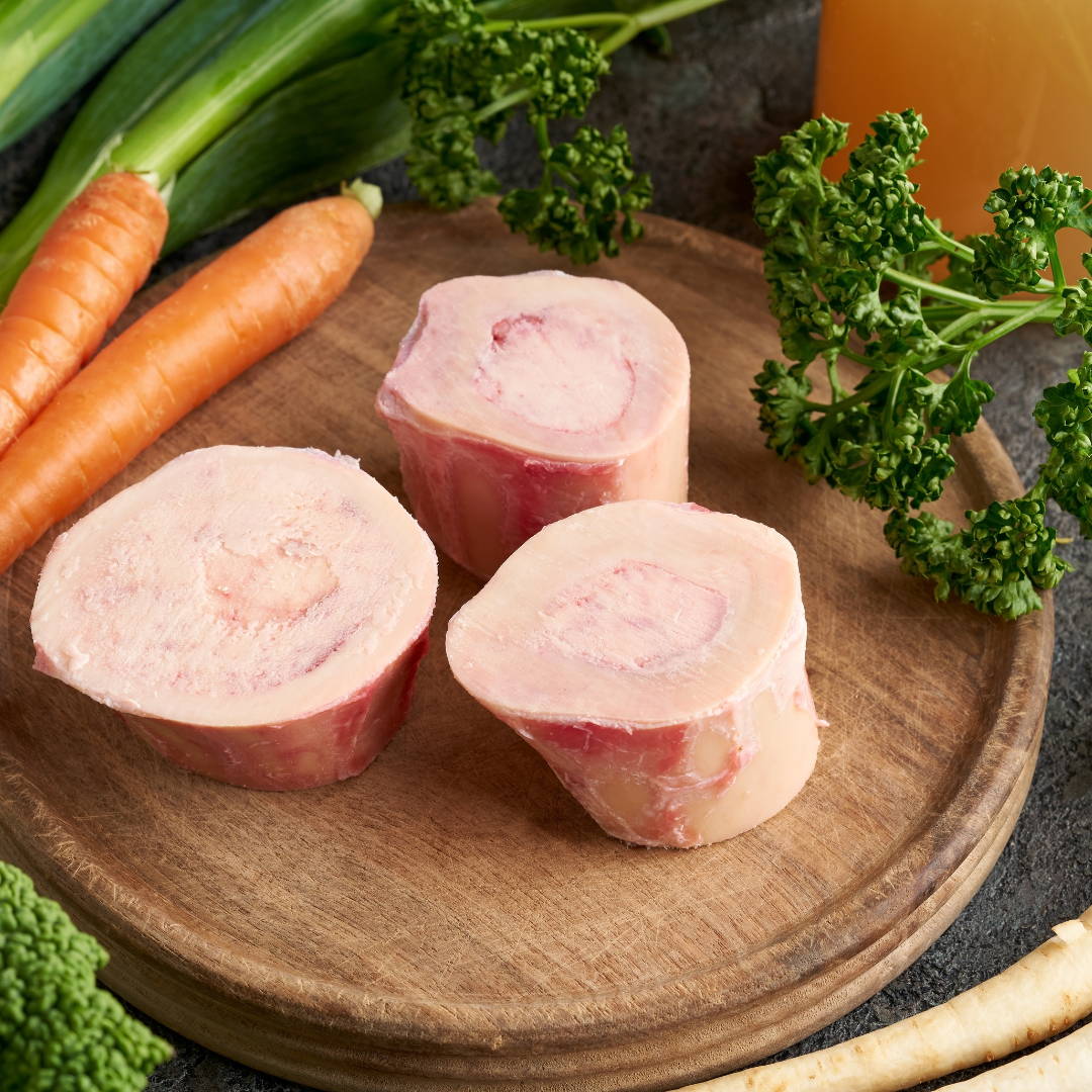 Raw marrow bones on circular wooden tray with carrots and parsley.