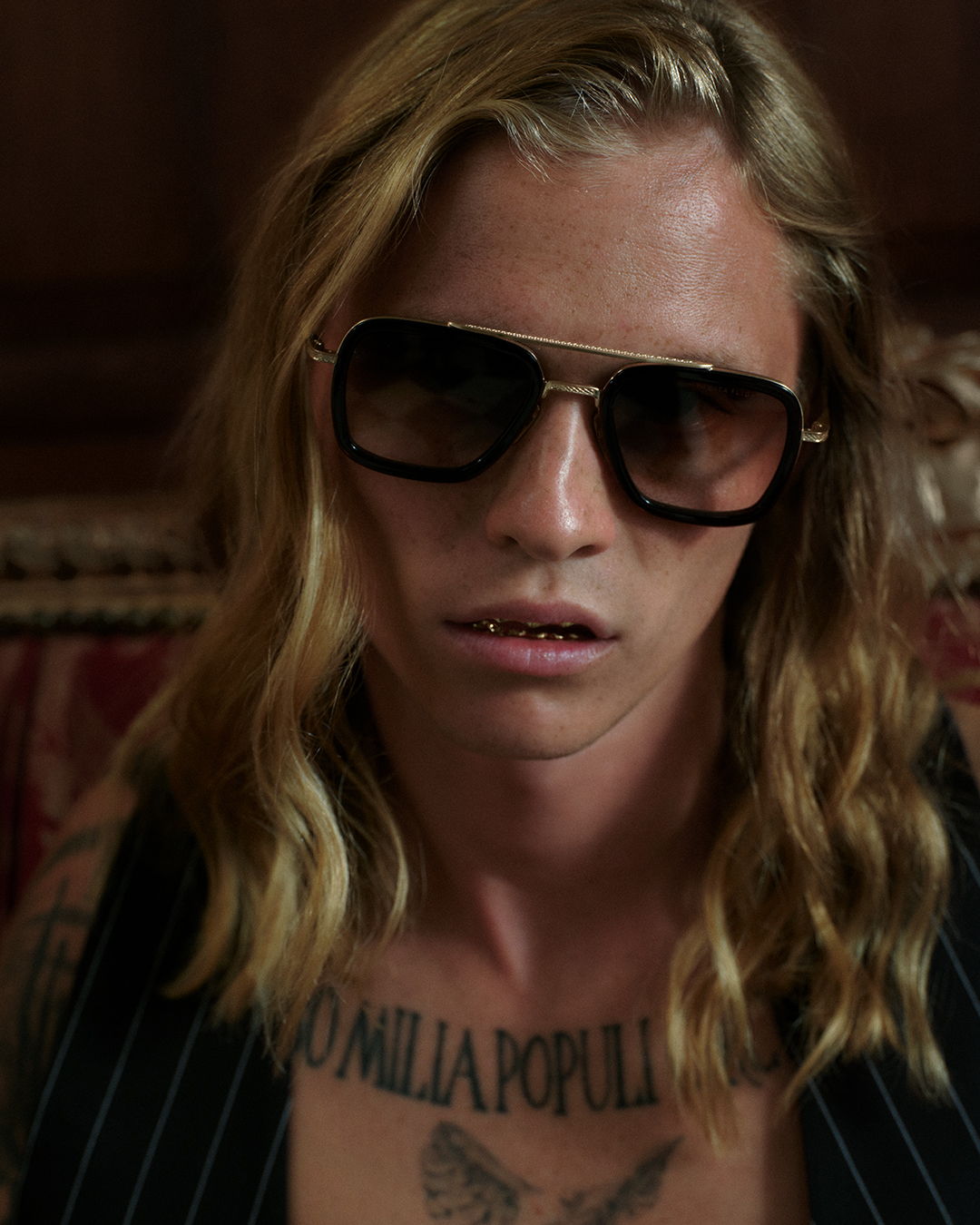 Person with long hair and sunglasses, sitting indoors with tattoos visible.
