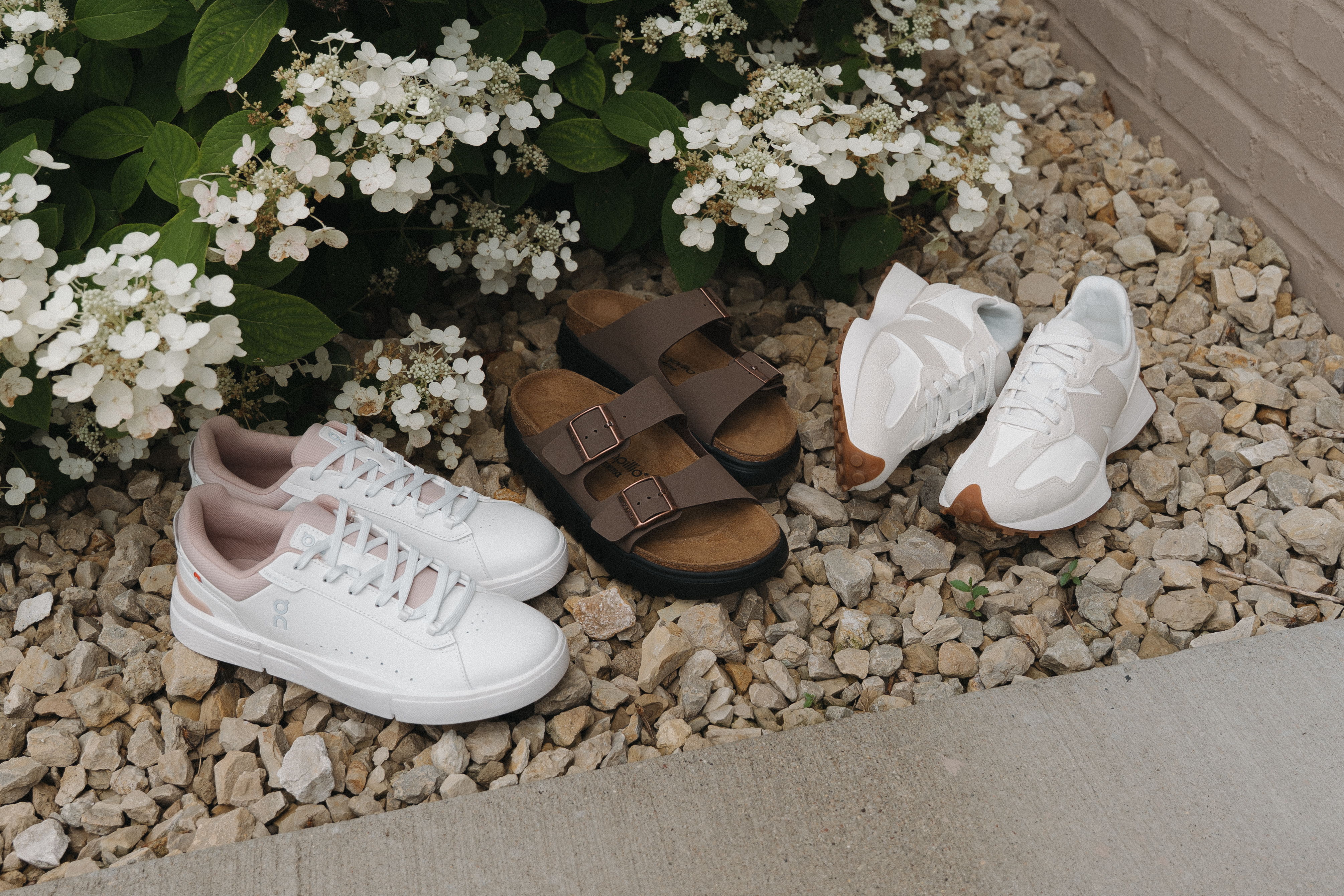 A variety of shoes arranged on gravel near flower bushes.