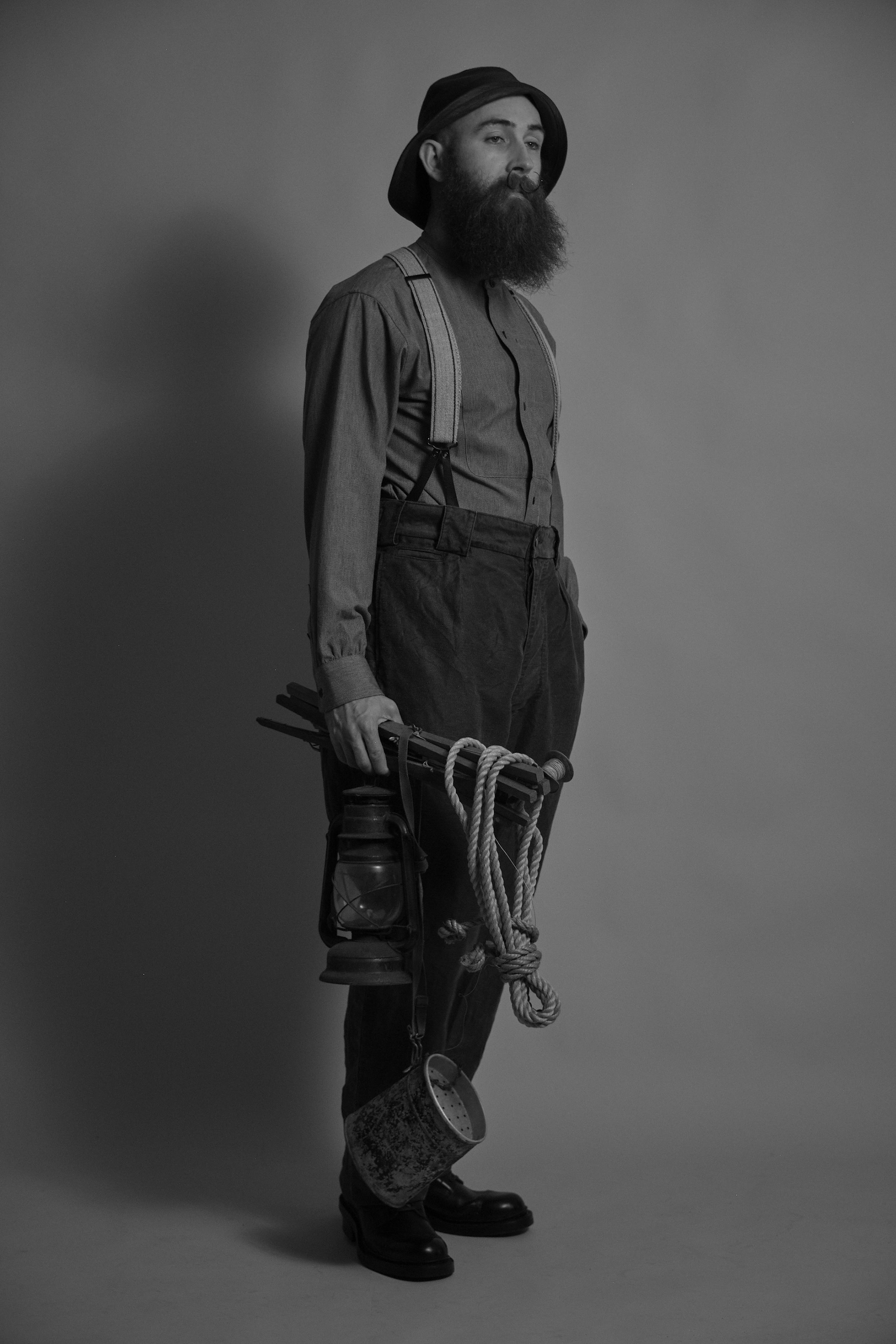 A man with a beard wearing suspenders, holding a lantern and rope.