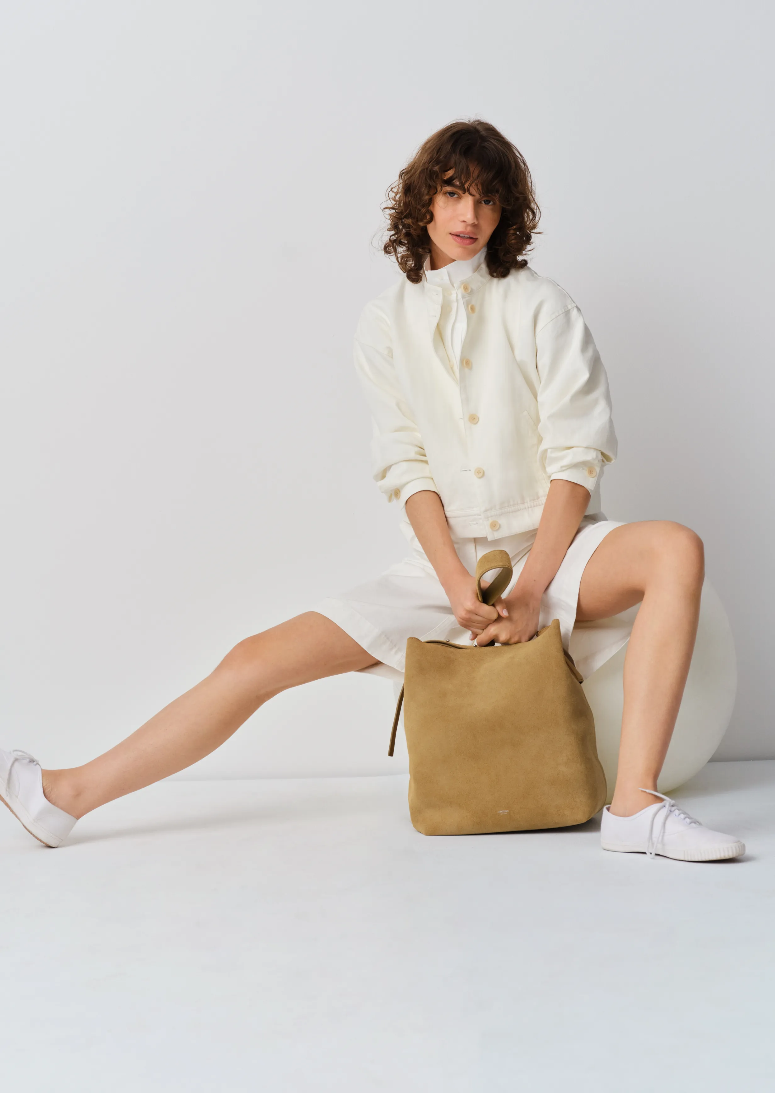 A person sitting with a tan bag, wearing a white outfit and sneakers.