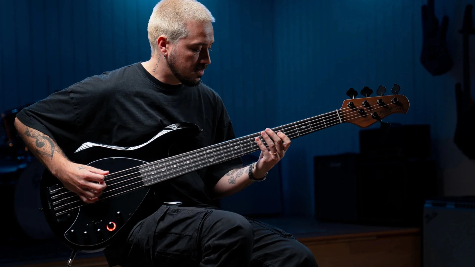 Person playing a Sterling by Music Man DarkRay 5 bass guitar in a dimly lit room.