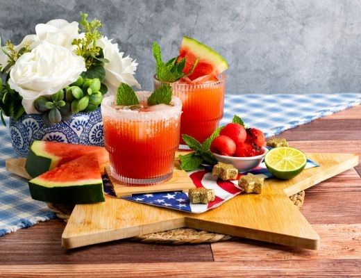 Two cocktails with watermelon and mint, surrounded by fruit and flowers on a table.