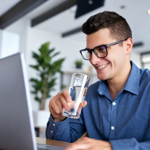 Man holding a glass of water while working on a laptop and smiling.