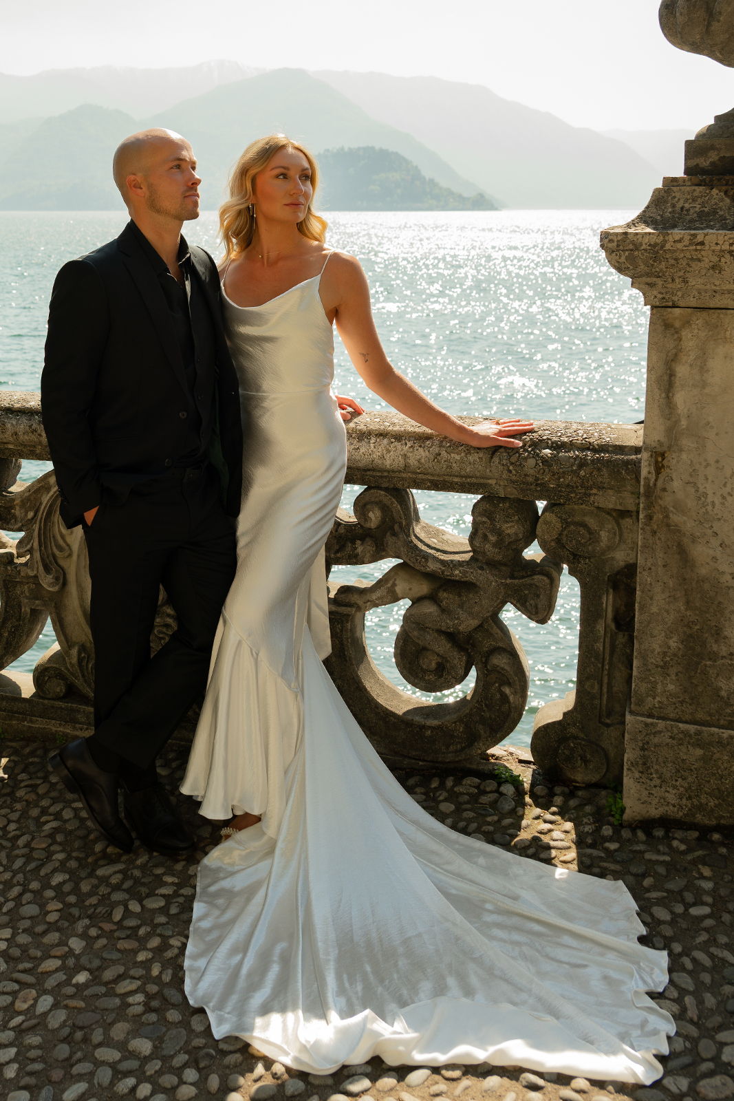 Couple in formal attire by a lake with mountains in the background.