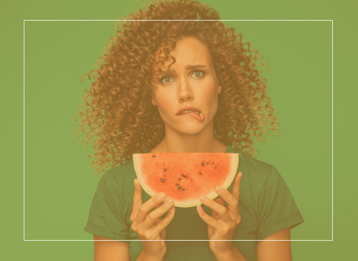 Woman holding up a juicy pink slice of watermelon and biting her lip – it could give her a tingly mouth if she has hay fever 