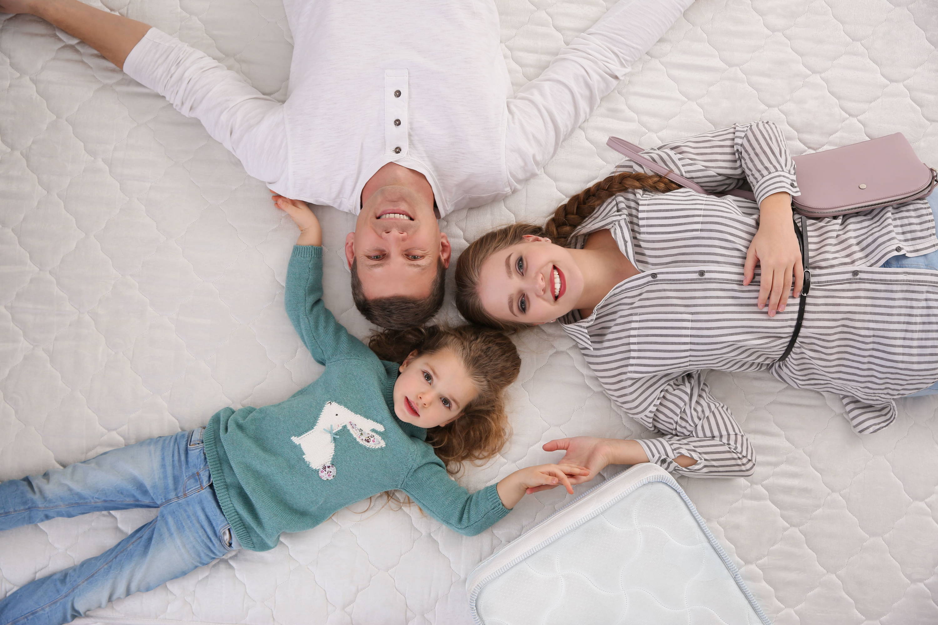 Overhead shot of a man and woman smiling and a child. All lie on a large mattress with heads touching in the centre of image.