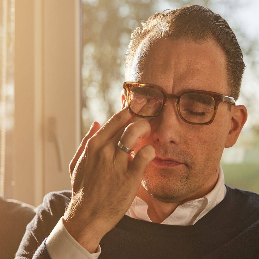  Muž si tře oko pod brýlemi, sedí doma na pohovce – alergie na roztoče může vést k zarudnutí, svědění a slzení očí