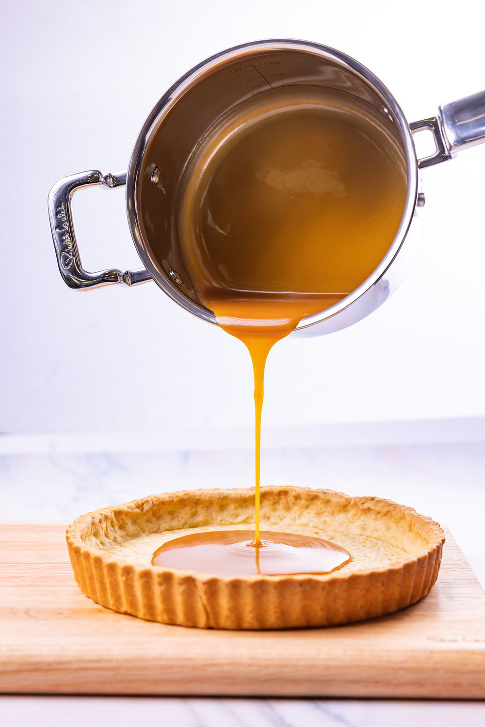A pot pouring caramel sauce into a tart shell on a wooden board.