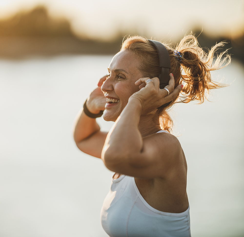 Woman enjoying music outdoors with headphones, smiling by the water.