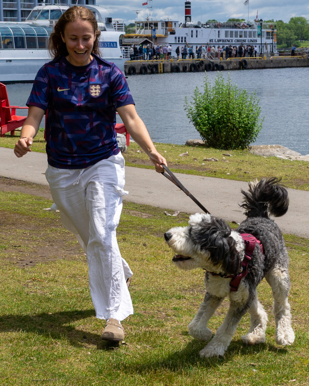 Person walking a dog near a waterfront on a sunny day.