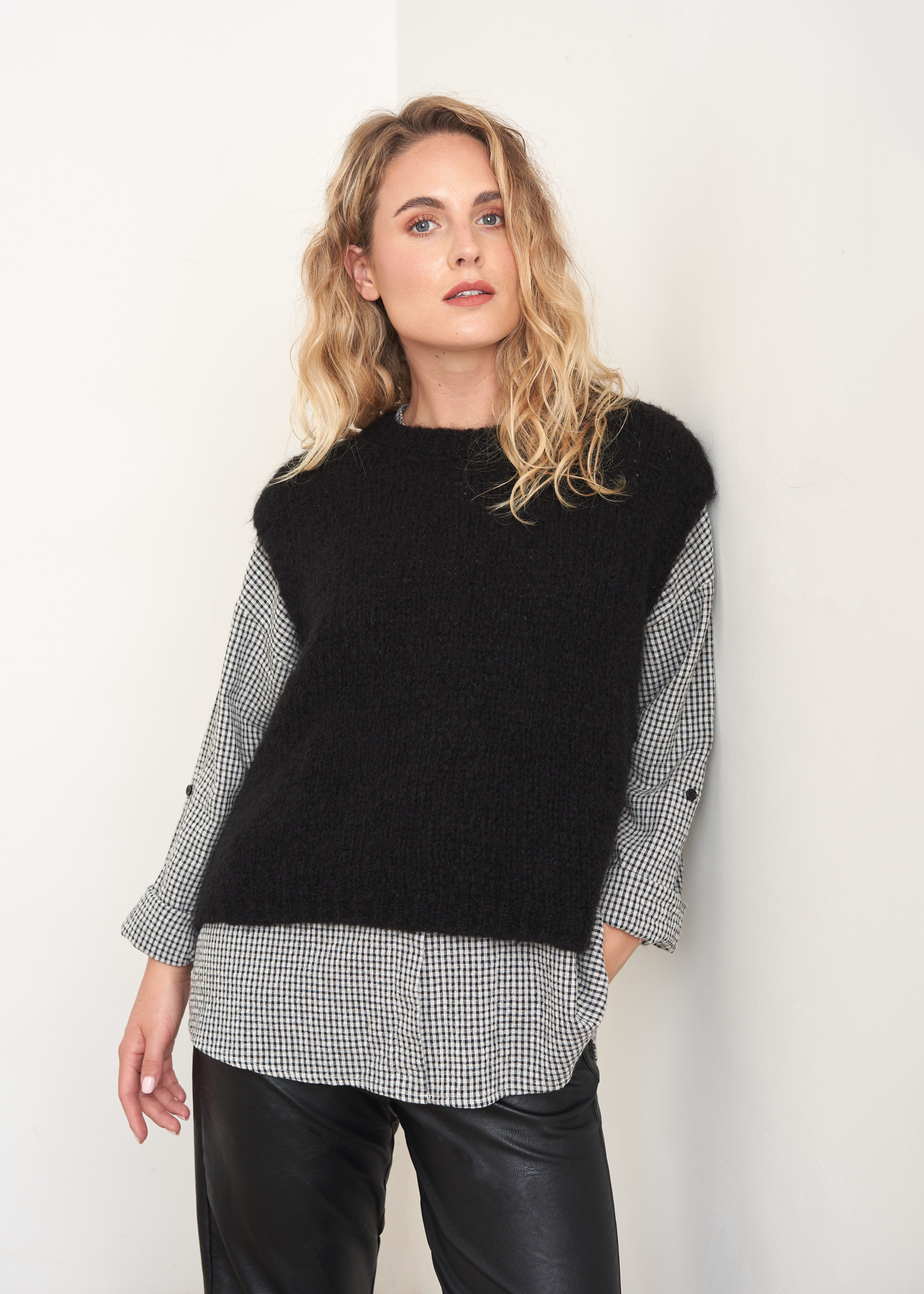A model wearing a black sleeveless knitted tank over a black and white checkered shirt and black leather trousers