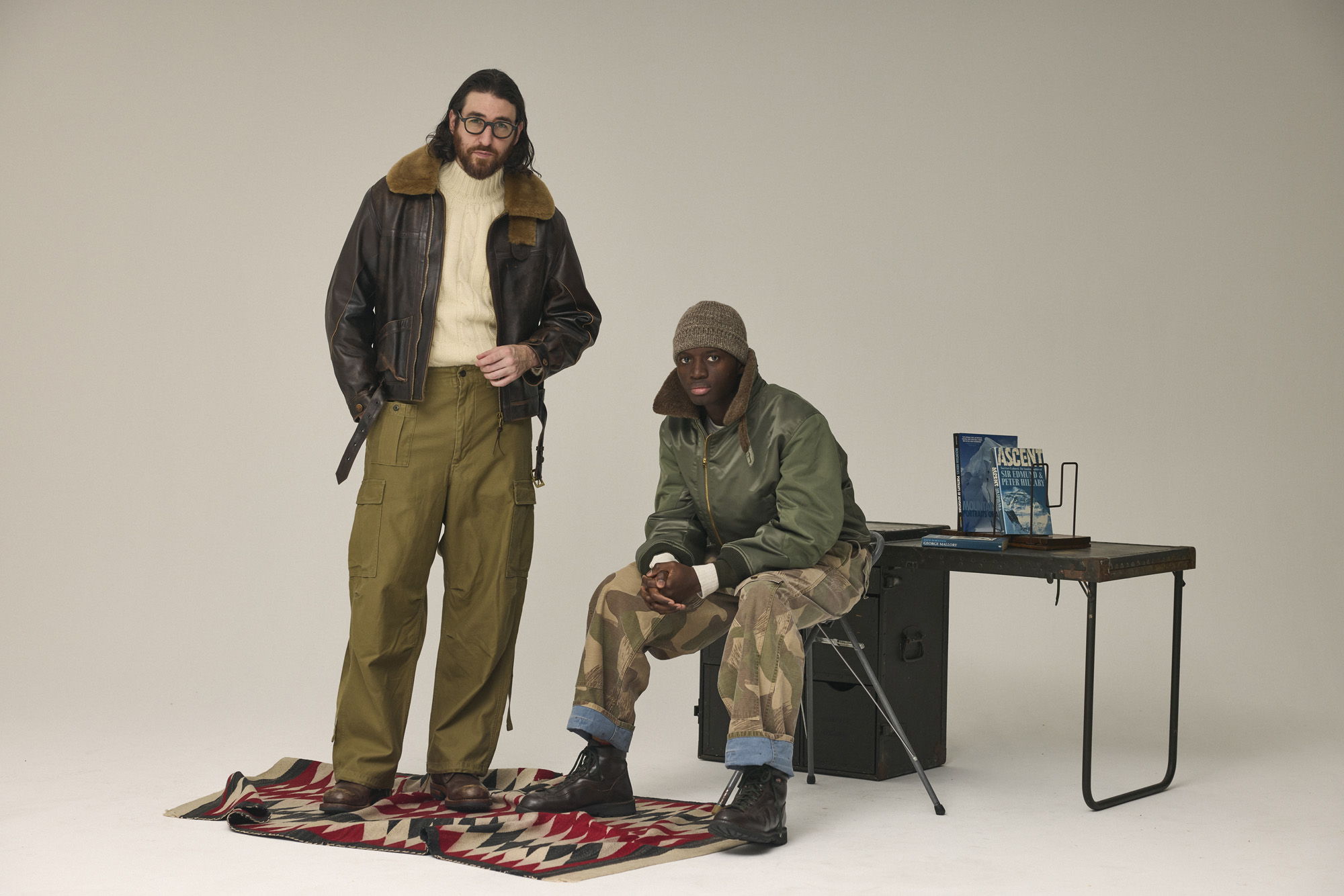 Two men in casual outfits, one standing and one seated, with a table nearby.