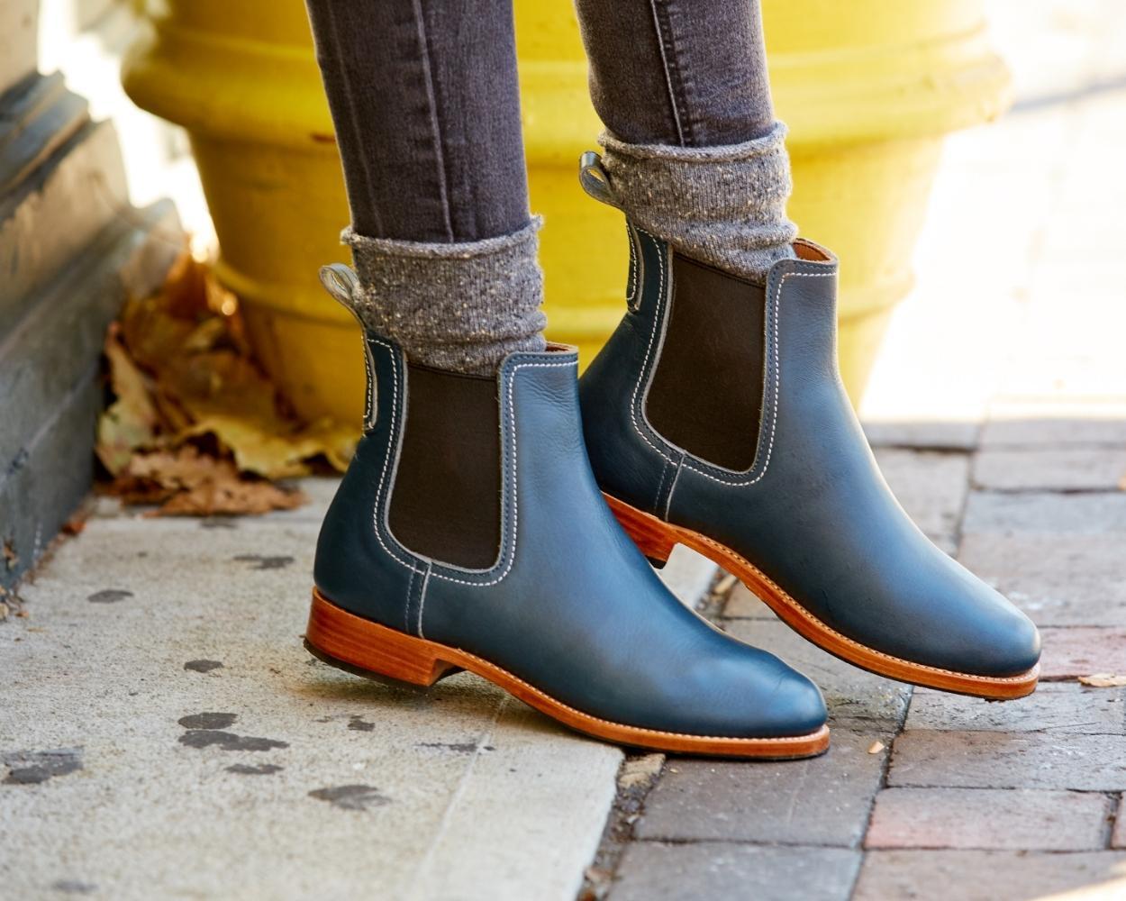 Close-up view of a person wearing the handcrafted Adelante Merida women's wide width leather Chelsea boots in denim blue, walking on a brick sidewalk.