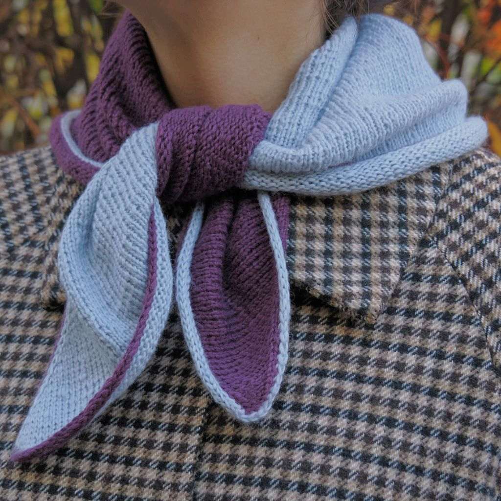 Eine Person, die ein blau-lila gestricktes Dreieckstuch über einem karierten Mantel trägt.