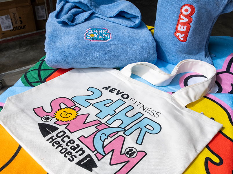 Blue folded hooded towles featuring 24hour swim and Revo patches, displayed beside a colourful tote bag printed with the bold Revo Fitness 24hr Ocean Heroes graphic, and sitting on a bold design sublimated beach towel