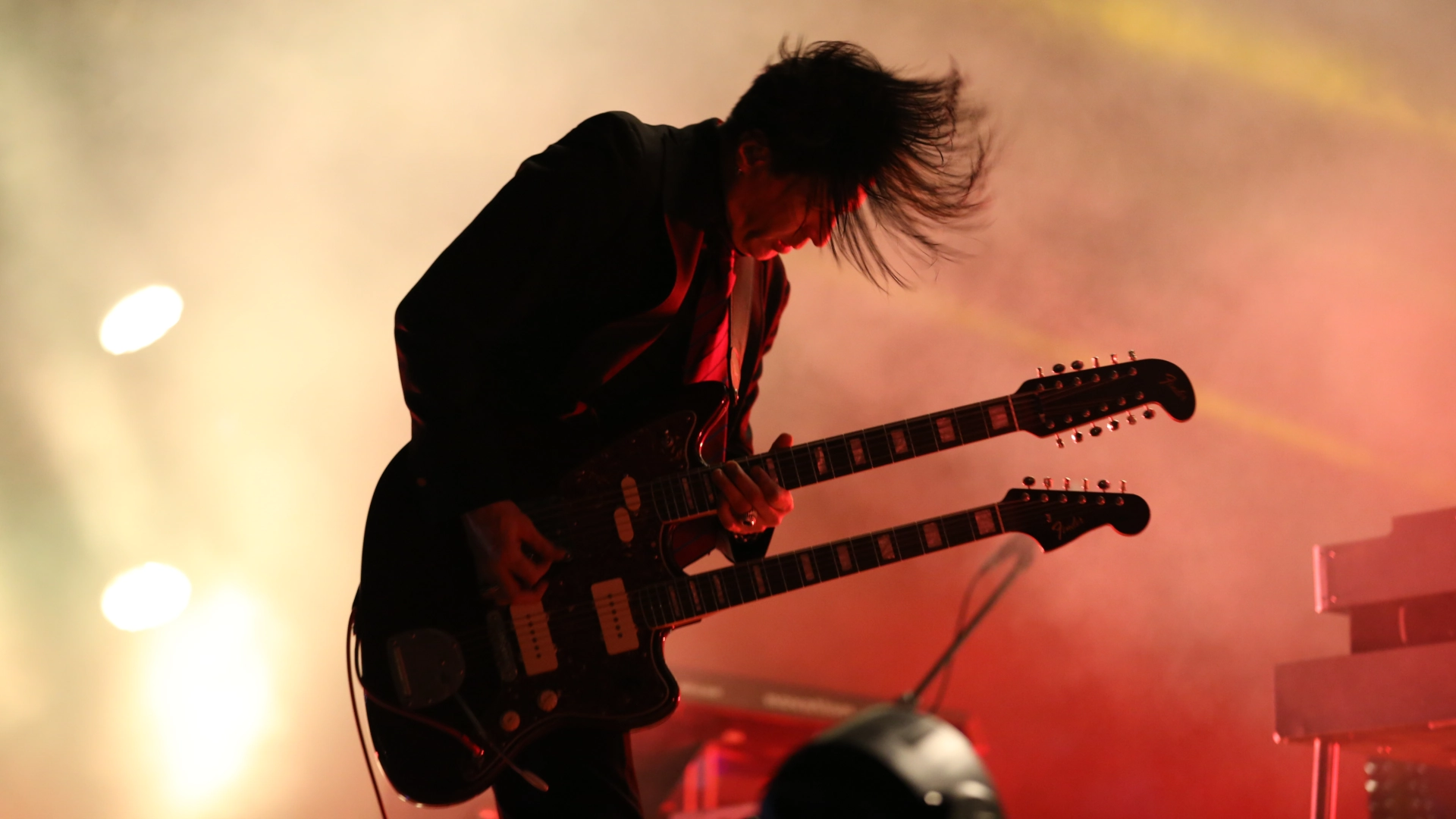 Silhouette of Troy Van Leeuwen playing a double neck Jazzmaster guitar in colourful lights on stage.