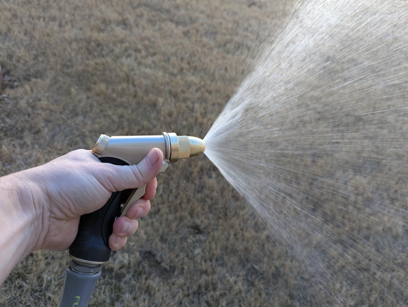 A hand holding a hose nozzle spraying water on dry grass.