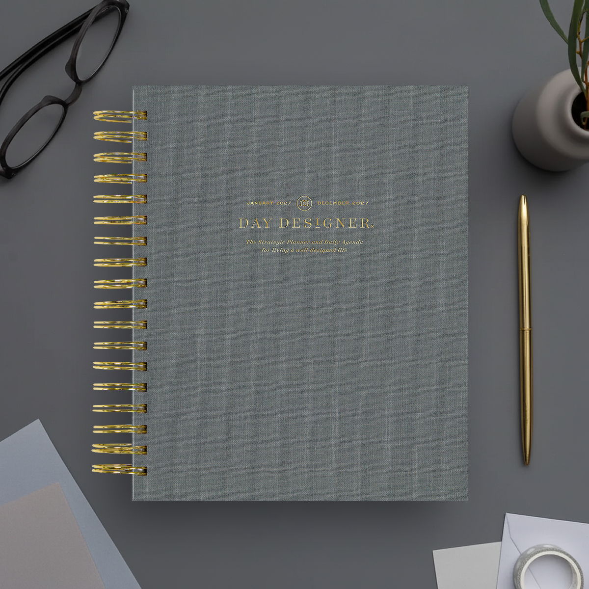 2027 day designer planner featuring charcoal bookcloth cover, gold binding and pen on a gray background