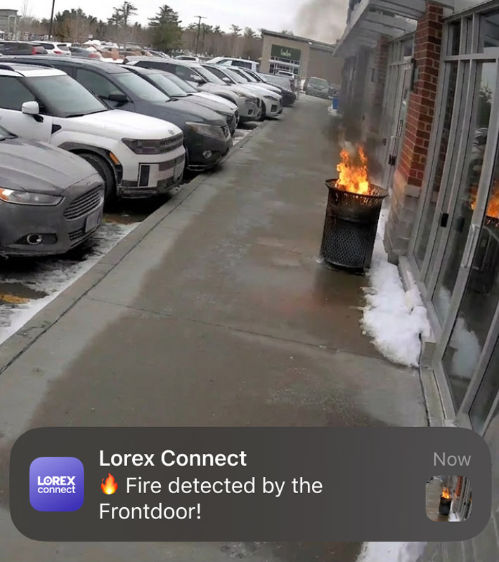 Security camera view of a fire in a trash bin outside a building with a mobile alert notification indicating fire detection