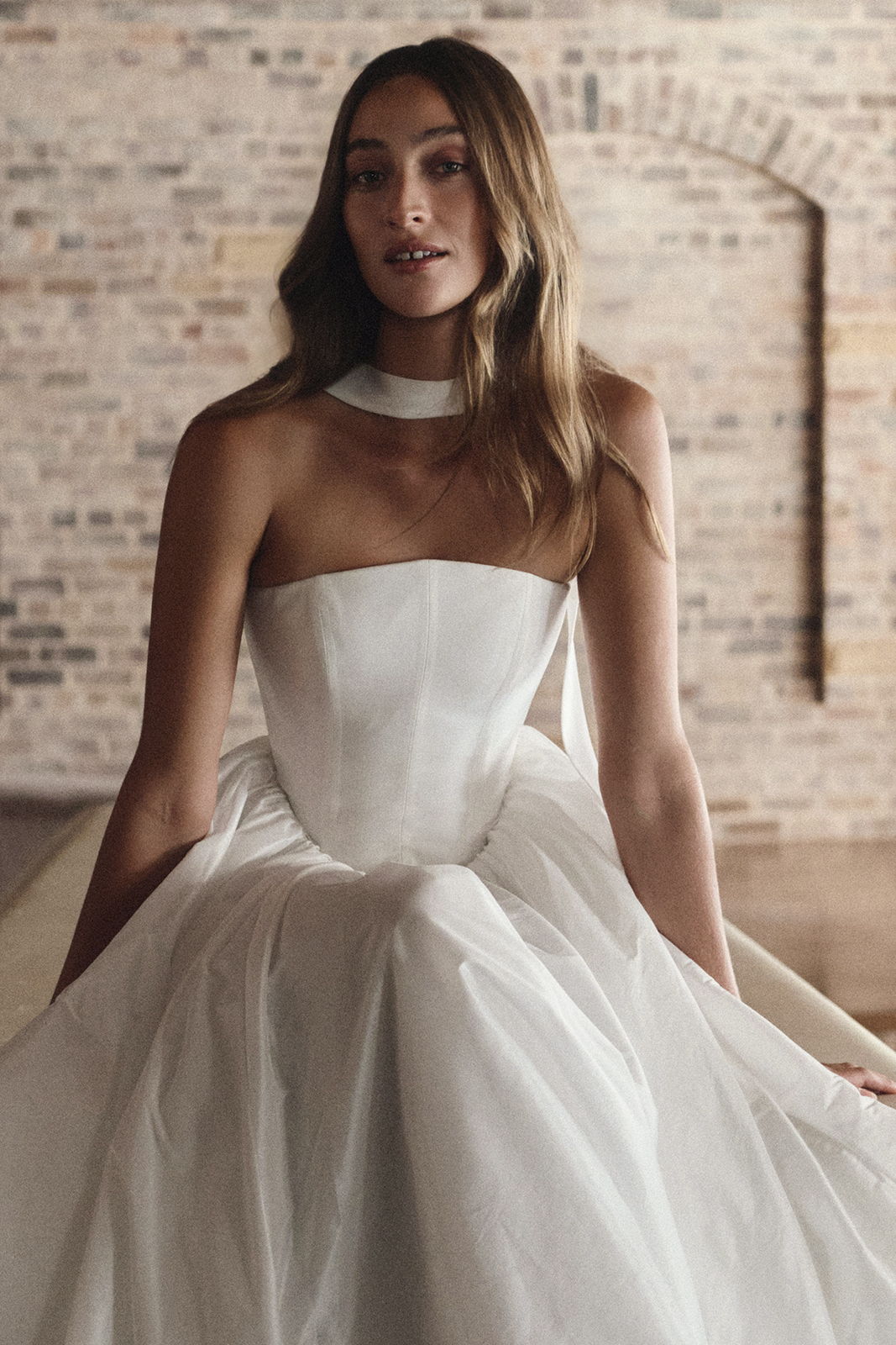 Woman in a strapless white dress sitting against a brick wall.