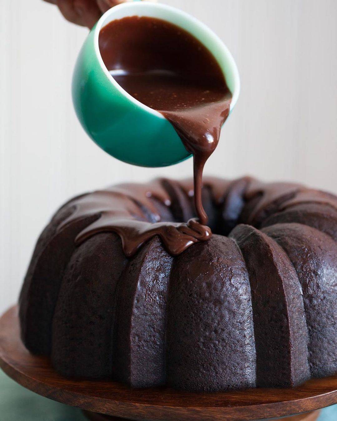 A delicious chocolate stout bundt cake ok a wooden cake stand, with chocolate glaze pouring out of a mug.