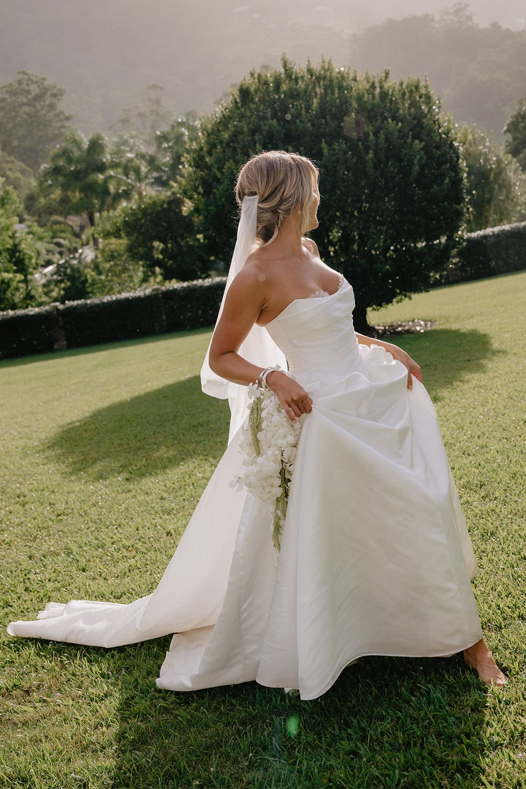 Bride in a white wedding dress holding her gown in a garden setting.