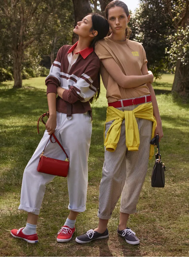 Two women in casual outfits pose together in a grassy outdoor setting.