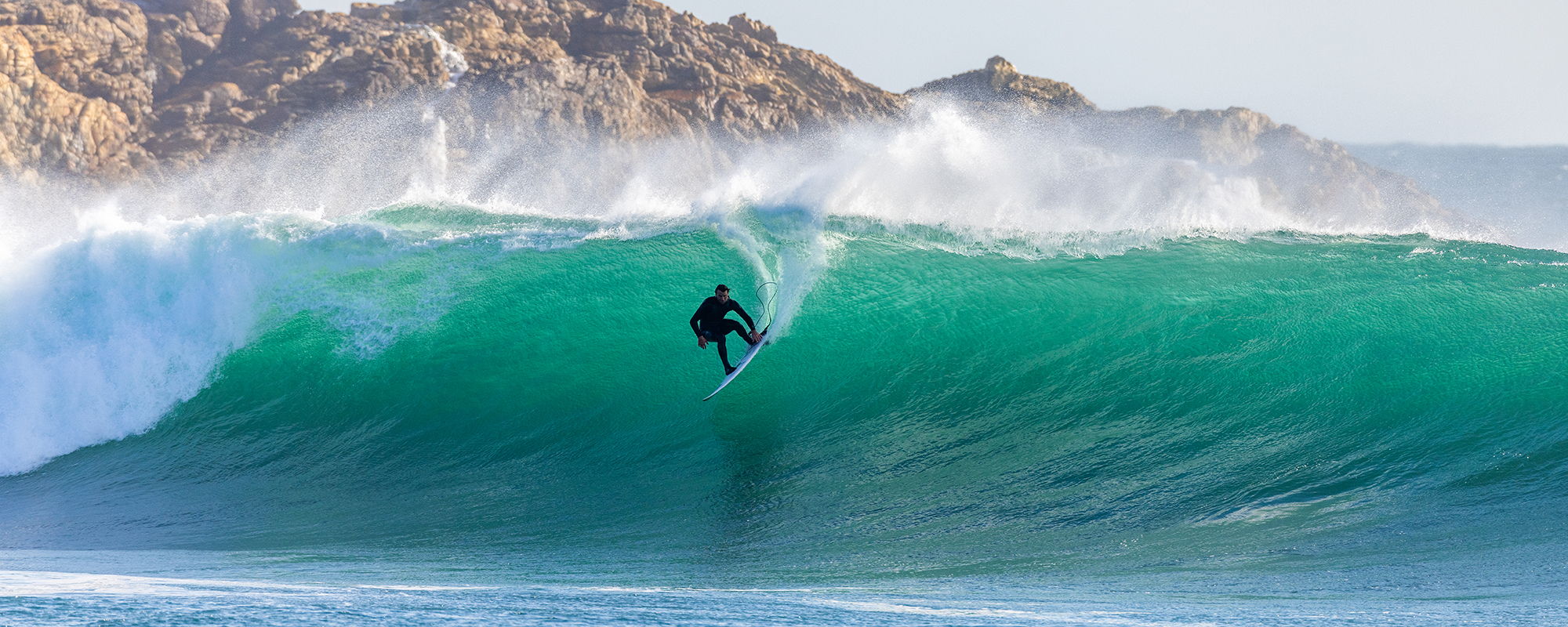 Un surfeur qui surf une grande vague turquoise, avec des falaises rocheuses en arrière-plan_Duotone_Ion Water