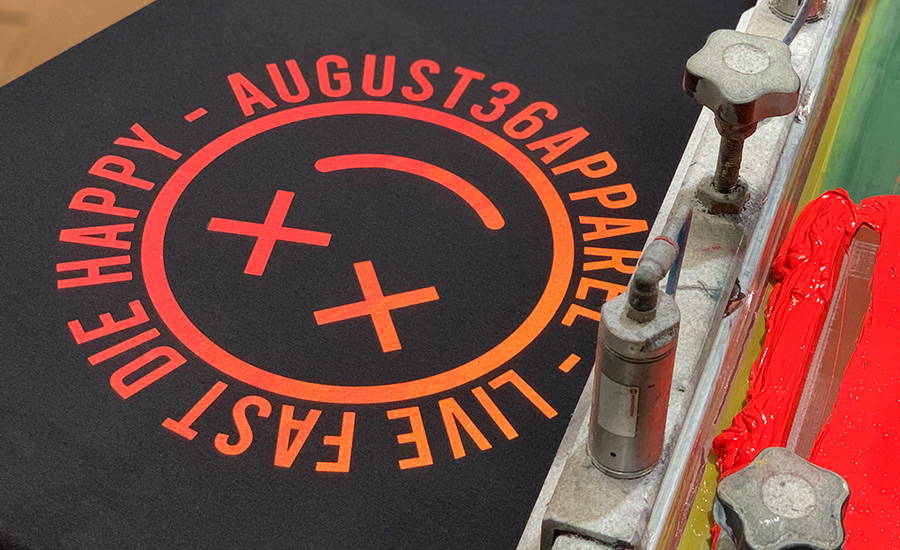 A specialty ink neon blend screen printed red and orange smiley face with x eyes on a black shirt. The shirt is still on the screen printing platen and the printing frame bracket is visible in the right corner showing bright red neon ink on the screen.