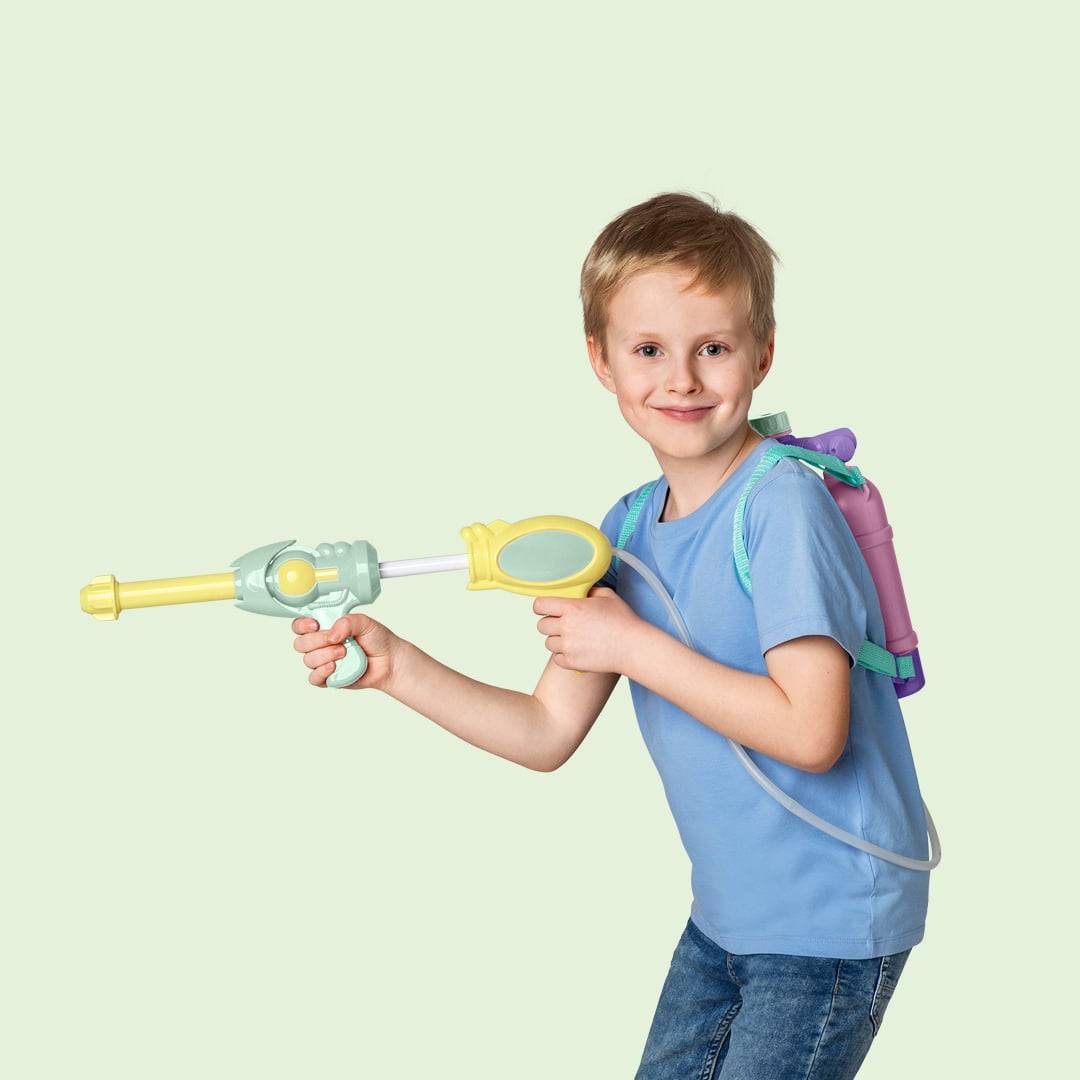Junge lächelt, während er einen gelben und grünen Wasserwerfer mit einem lila Rucksack vor einem hellgrünen Hintergrund hält.