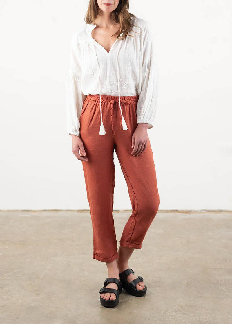 A model wearing a pear of terracotta coloured tapered linen trousers with black chunky buckle slides and a white bohemian inspired off white blouse with tassel detailing