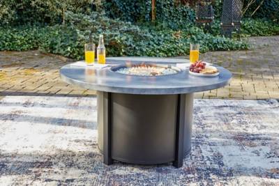 A fire pit table sits on a grey concrete patio. A fire is burning in the middle of the table with bottles and glasses filled with orange juice and drinks. A plate of fruit also sits on the table.