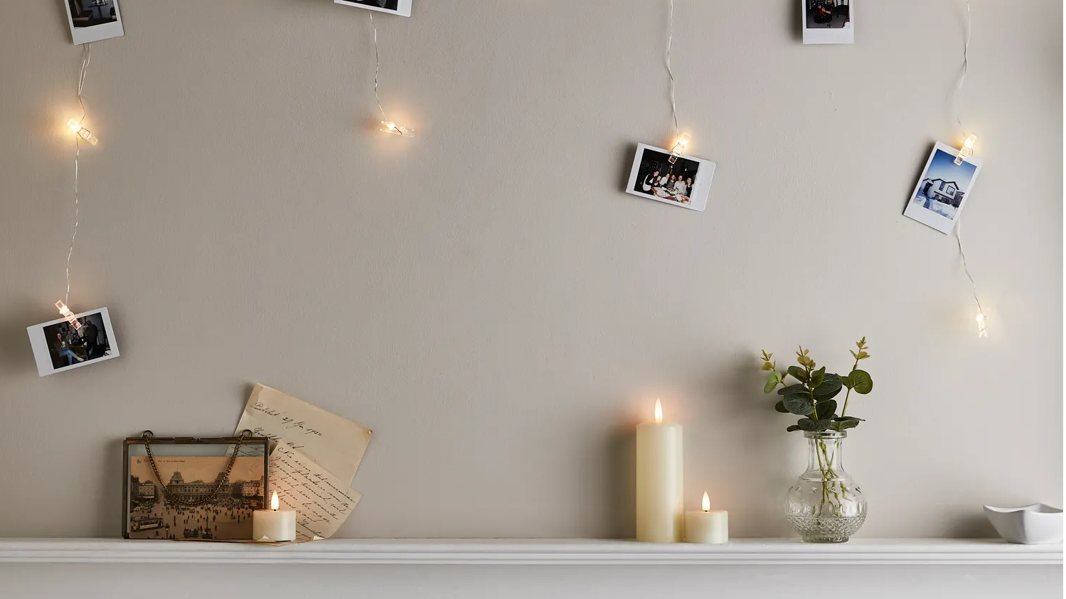 Slim ivory pillar candle and votive candles sat on mantel with peg fairy lights illuminated with polaroids attached 