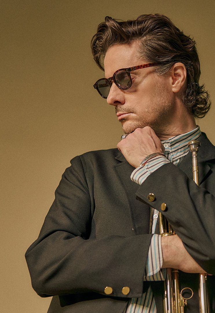 Man in sunglasses holding a trumpet, wearing a stylish suit against a plain background.