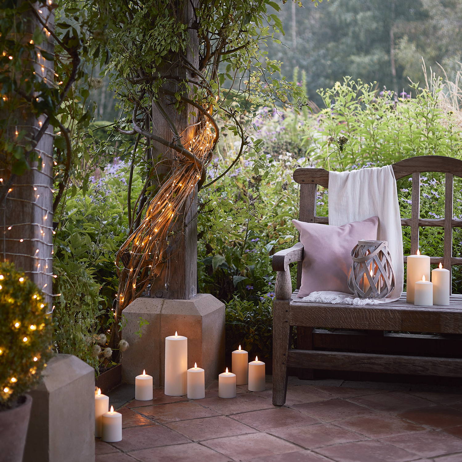 Jardin avec banc sur lequel se trouve une lanterne et des bougies avec des bougies sur le sol et des guirlandes lumineuses.