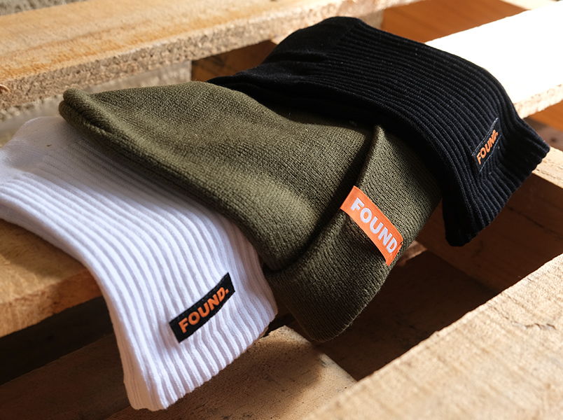 Stack of ribbed socks and beanies in olive, black and white featuring an orange 