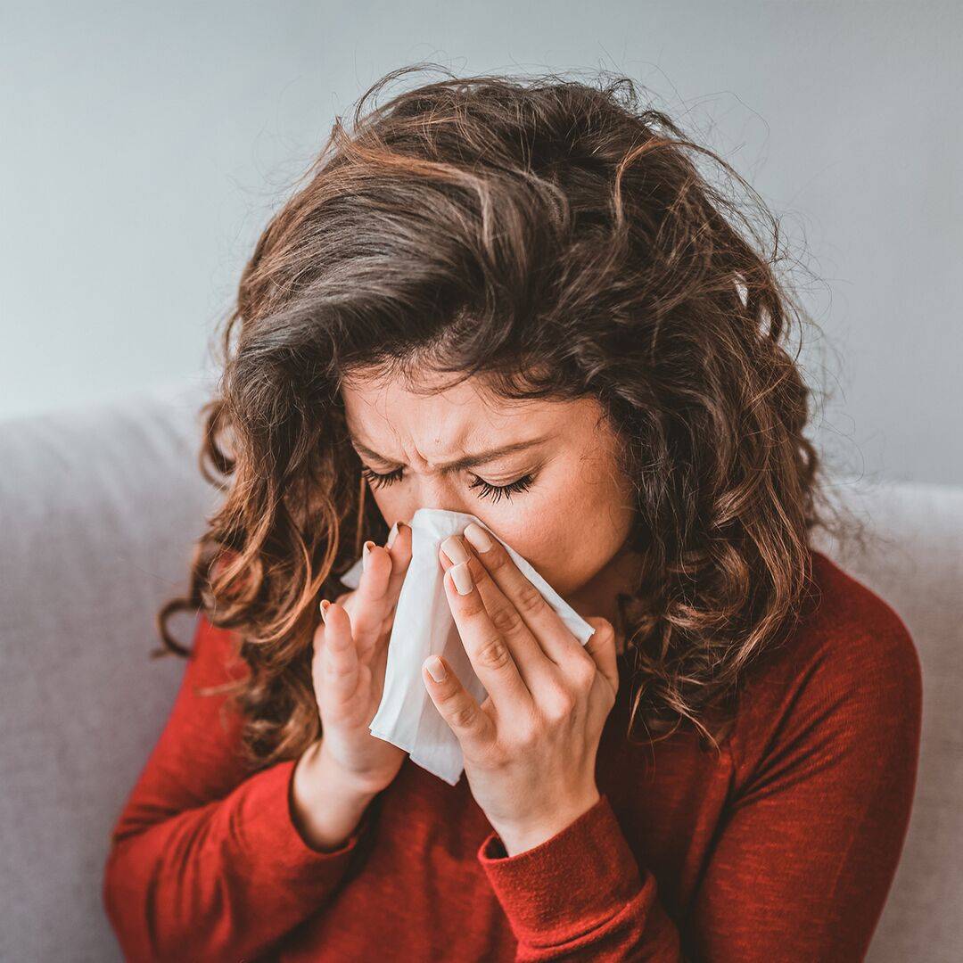Une femme brune se penche en avant et se mouche. En cas d’allergies, il est possible d’avoir le nez bouché et qui coule en même temps.