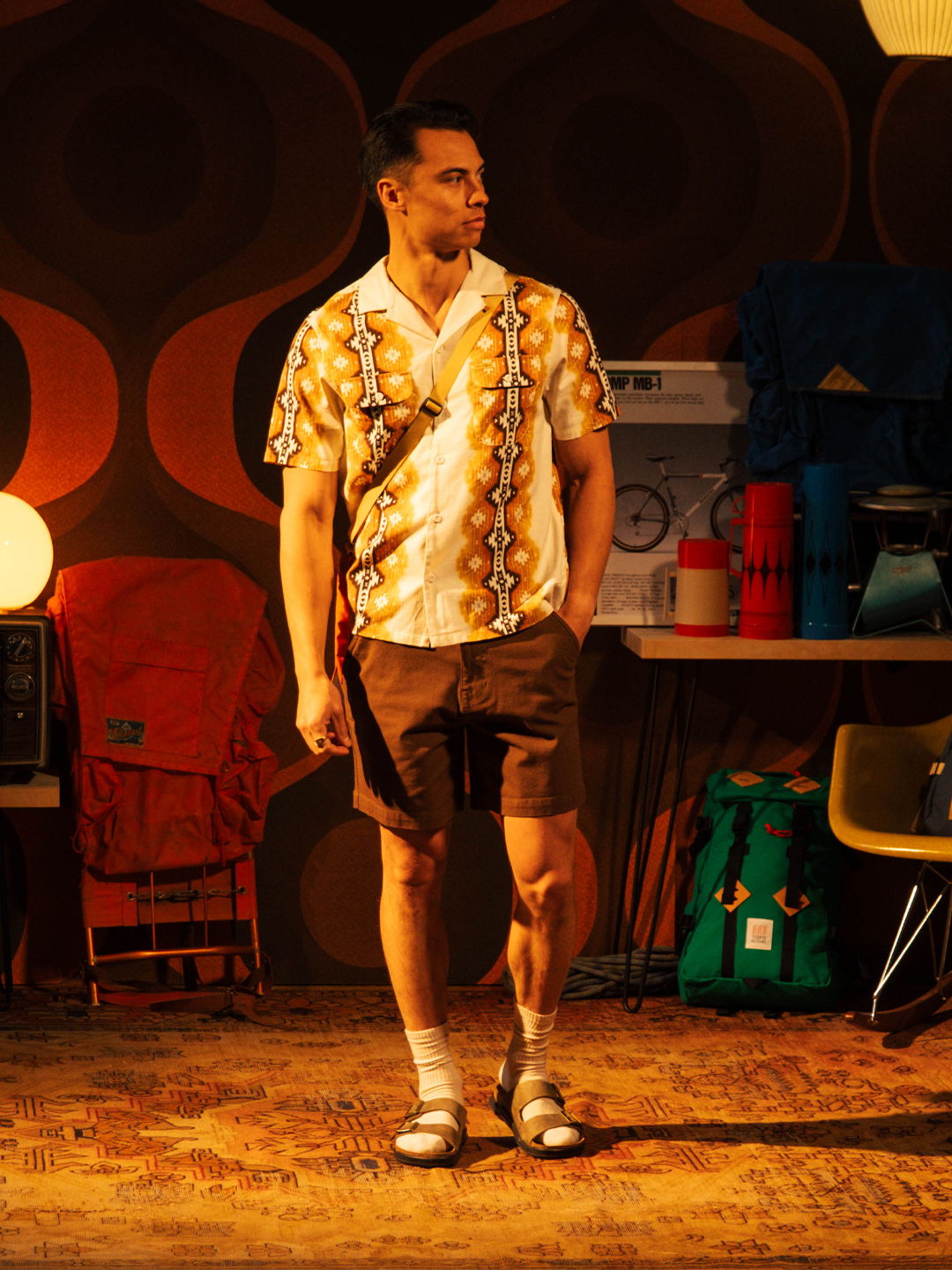 Man in patterned shirt and shorts, standing in a retro-themed room.