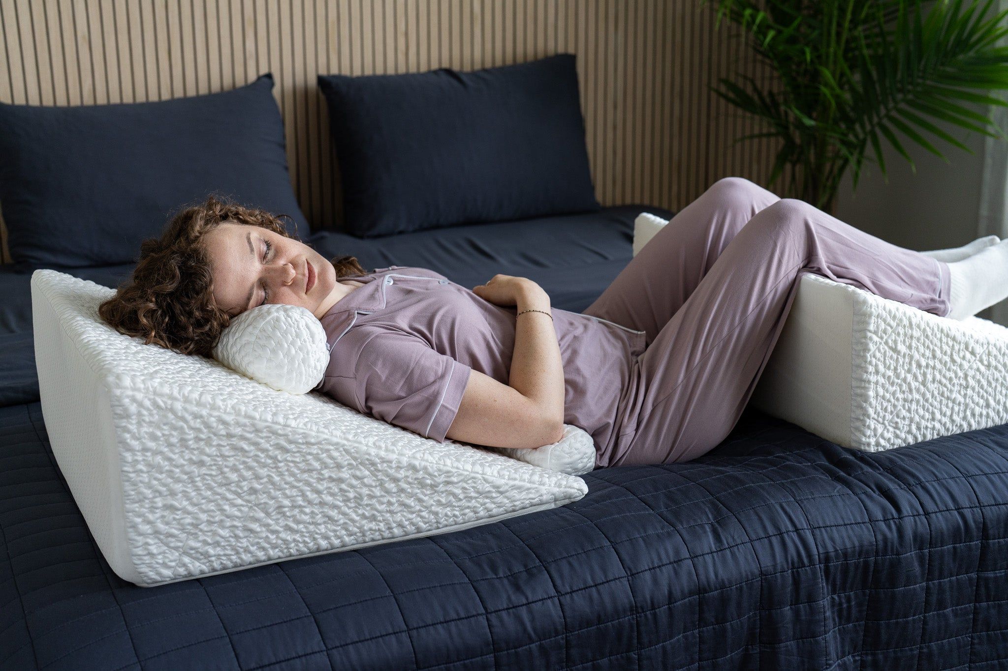 Person lying on a bed with a wedge pillow for neck support and another for leg support