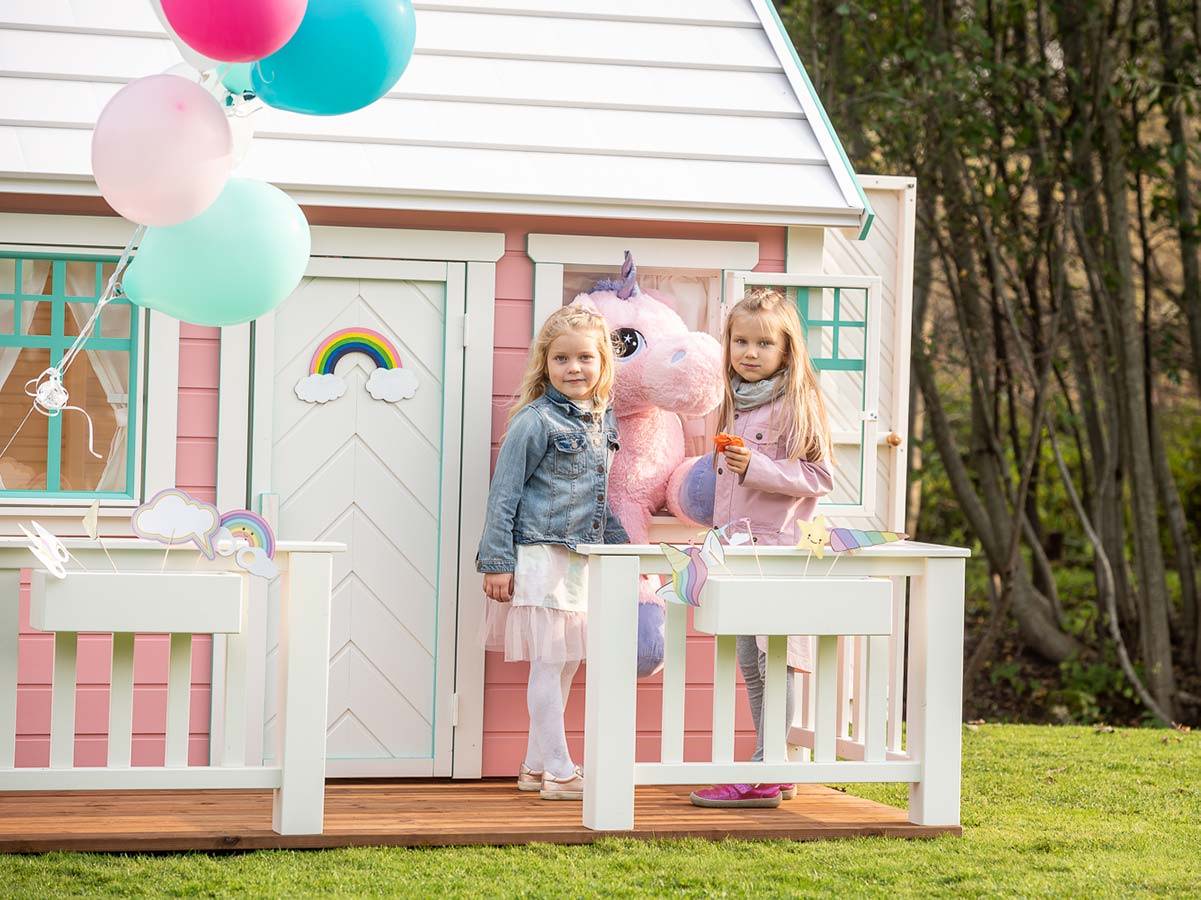 Two girls are standing on the wooden terrace of a pink Outdoor Playhouse by WholeWoodPlayhouses