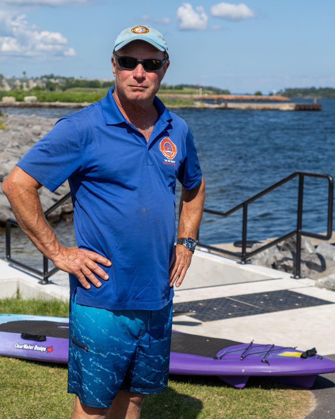 Man wearing sunglasses and a blue shirt stands by the water.