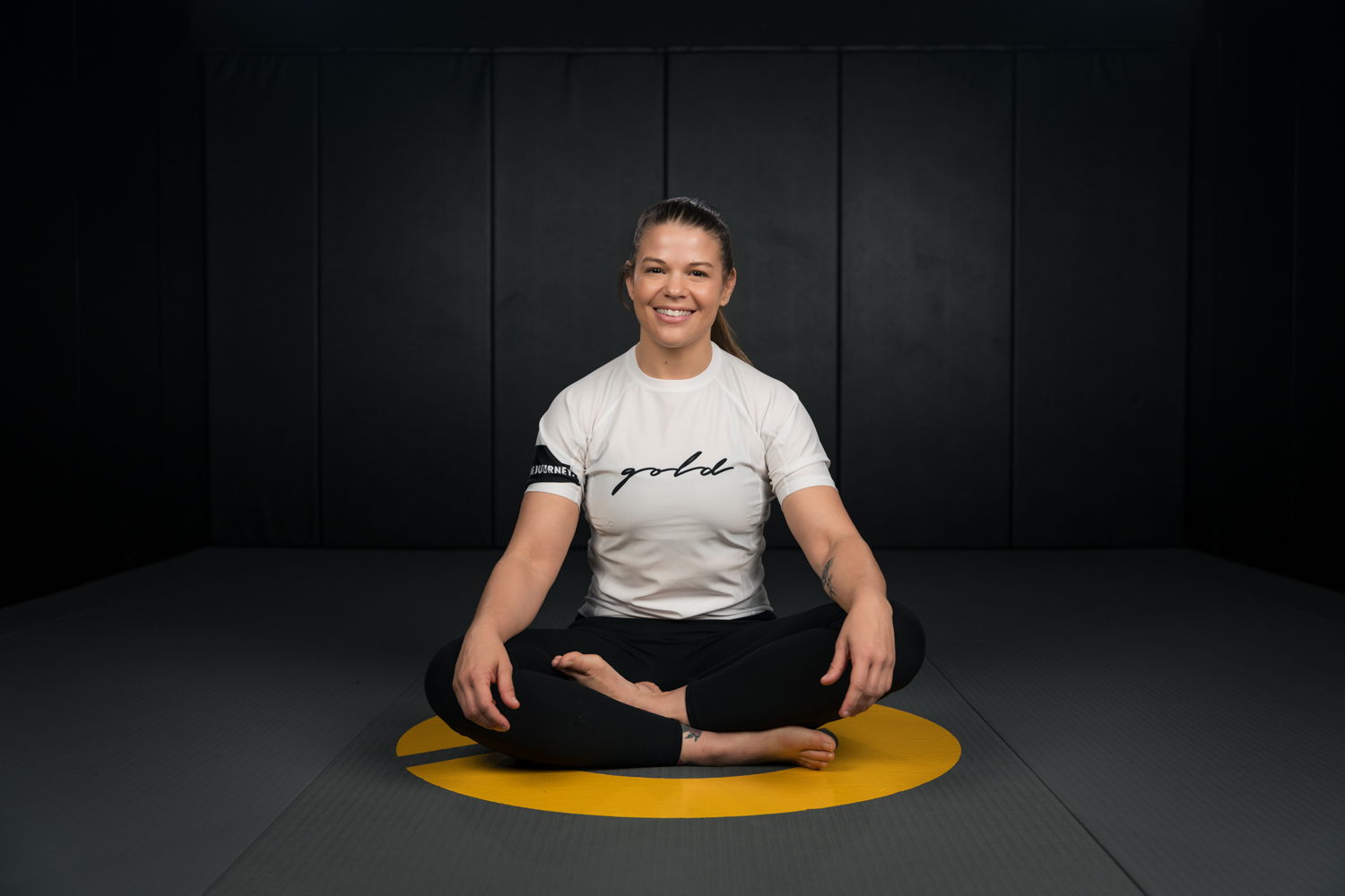 Person sitting cross-legged on a mat, smiling, with a dark background.