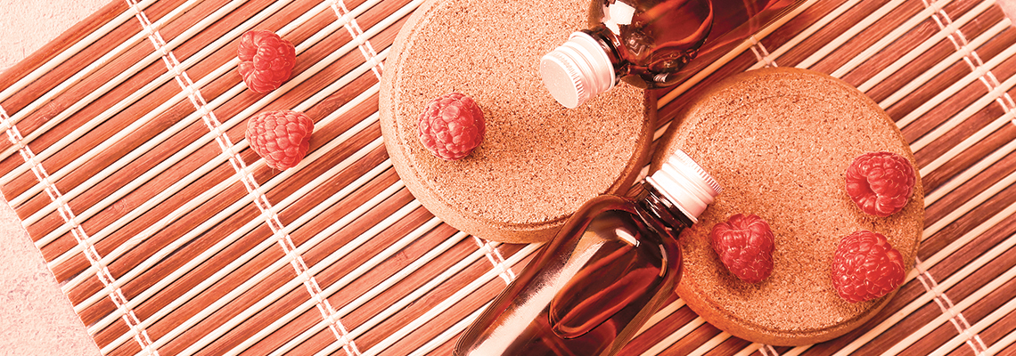 Two round wooden coasters with essential oil bottles and raspberries on a bamboo mat.
