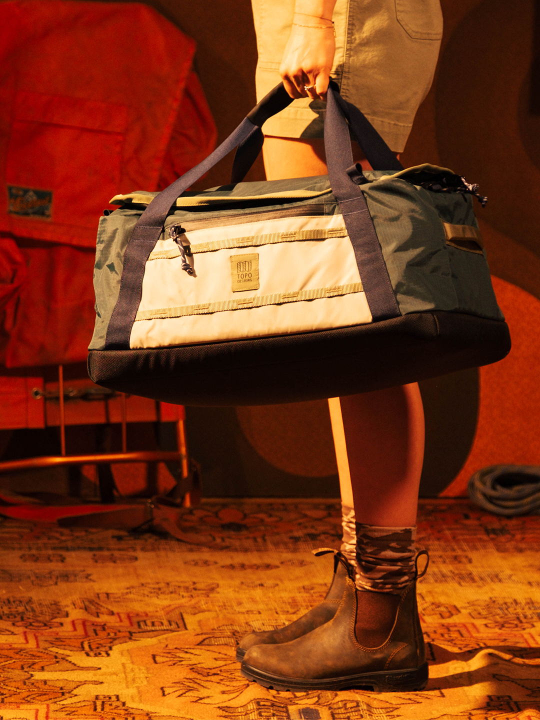Person holding a duffel bag, wearing boots, with camping gear in background.