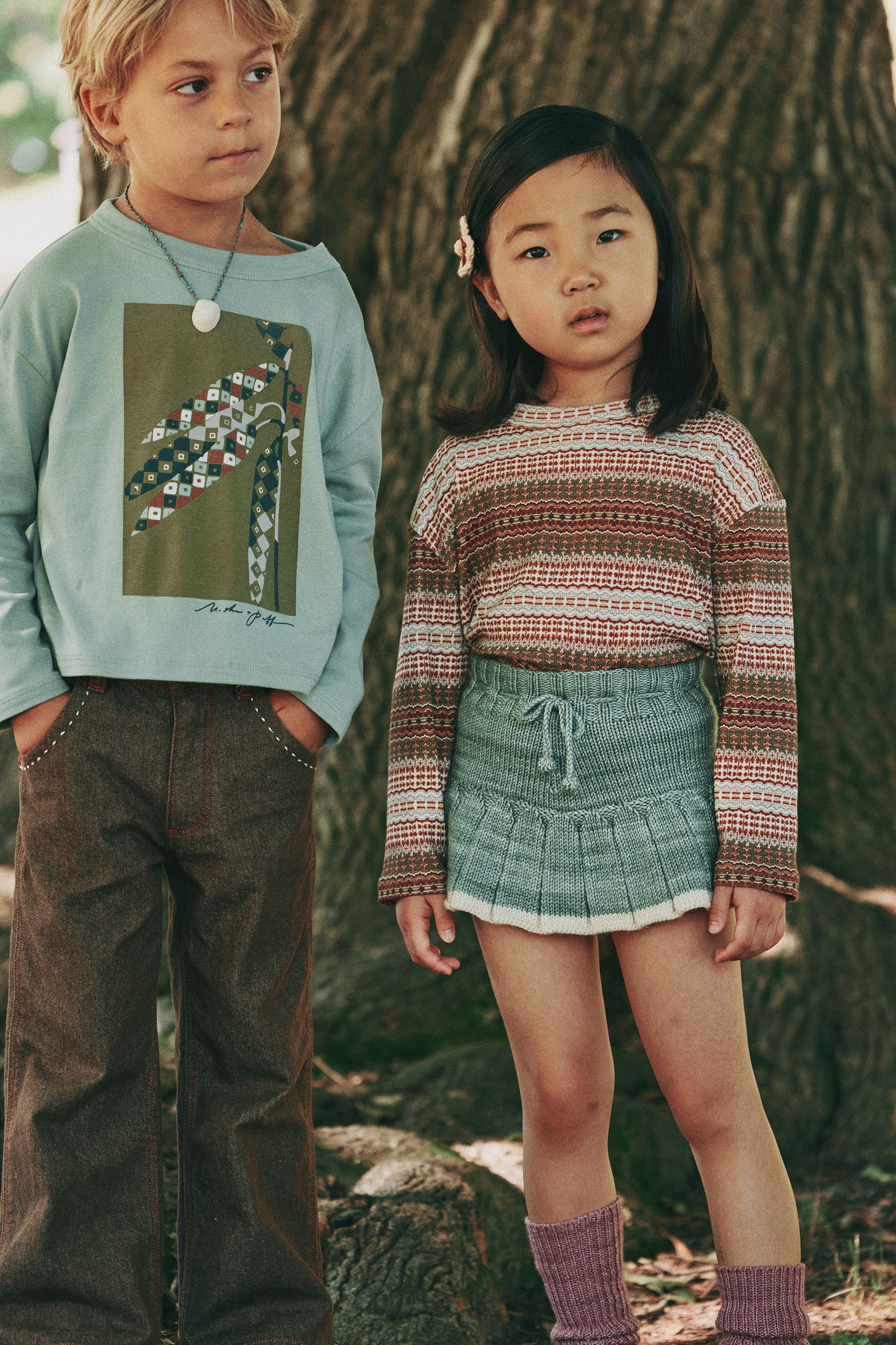 Two children standing near a tree, wearing casual clothing.