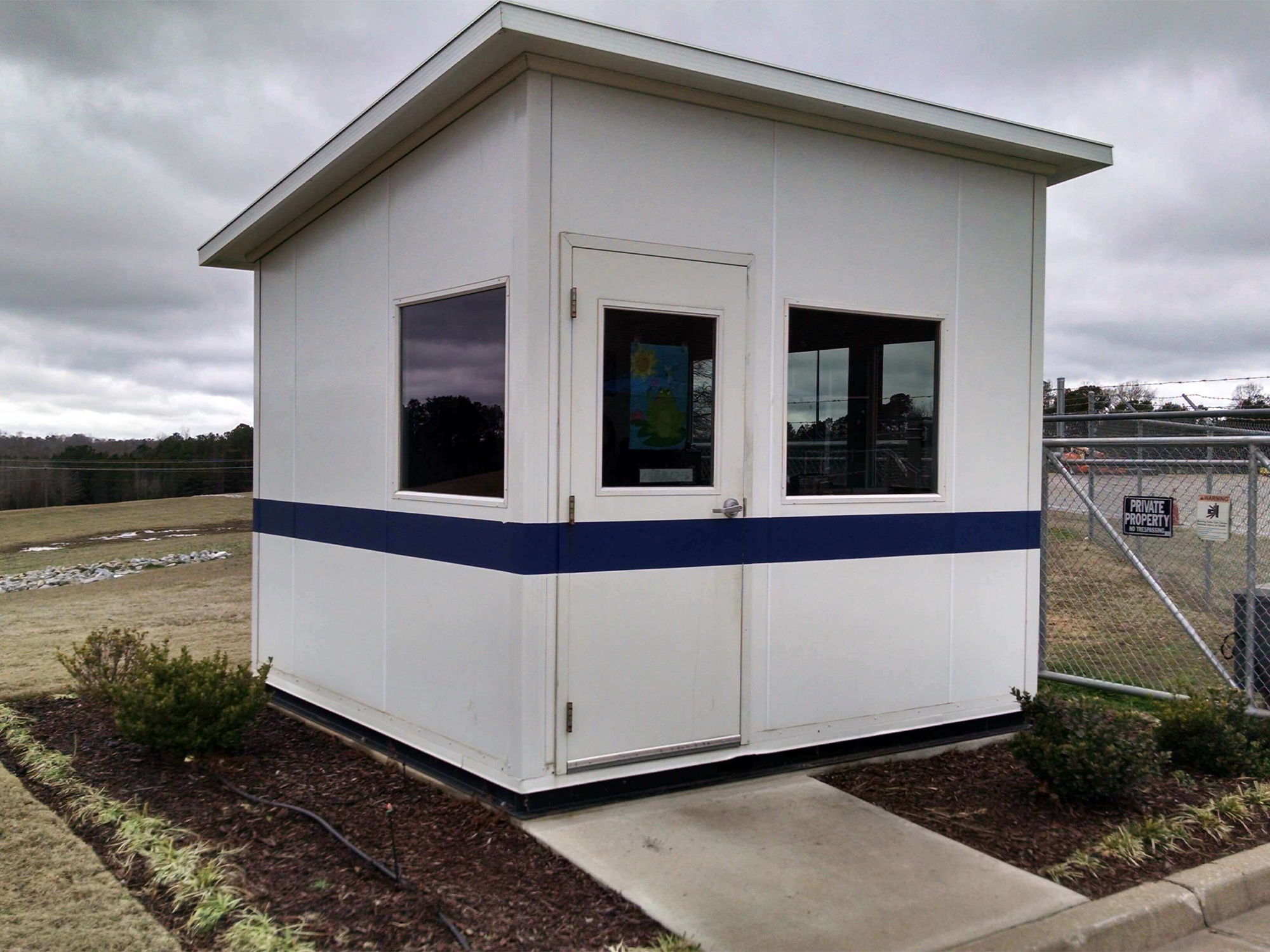 Small white security booth with blue stripe and surrounding landscaping.
