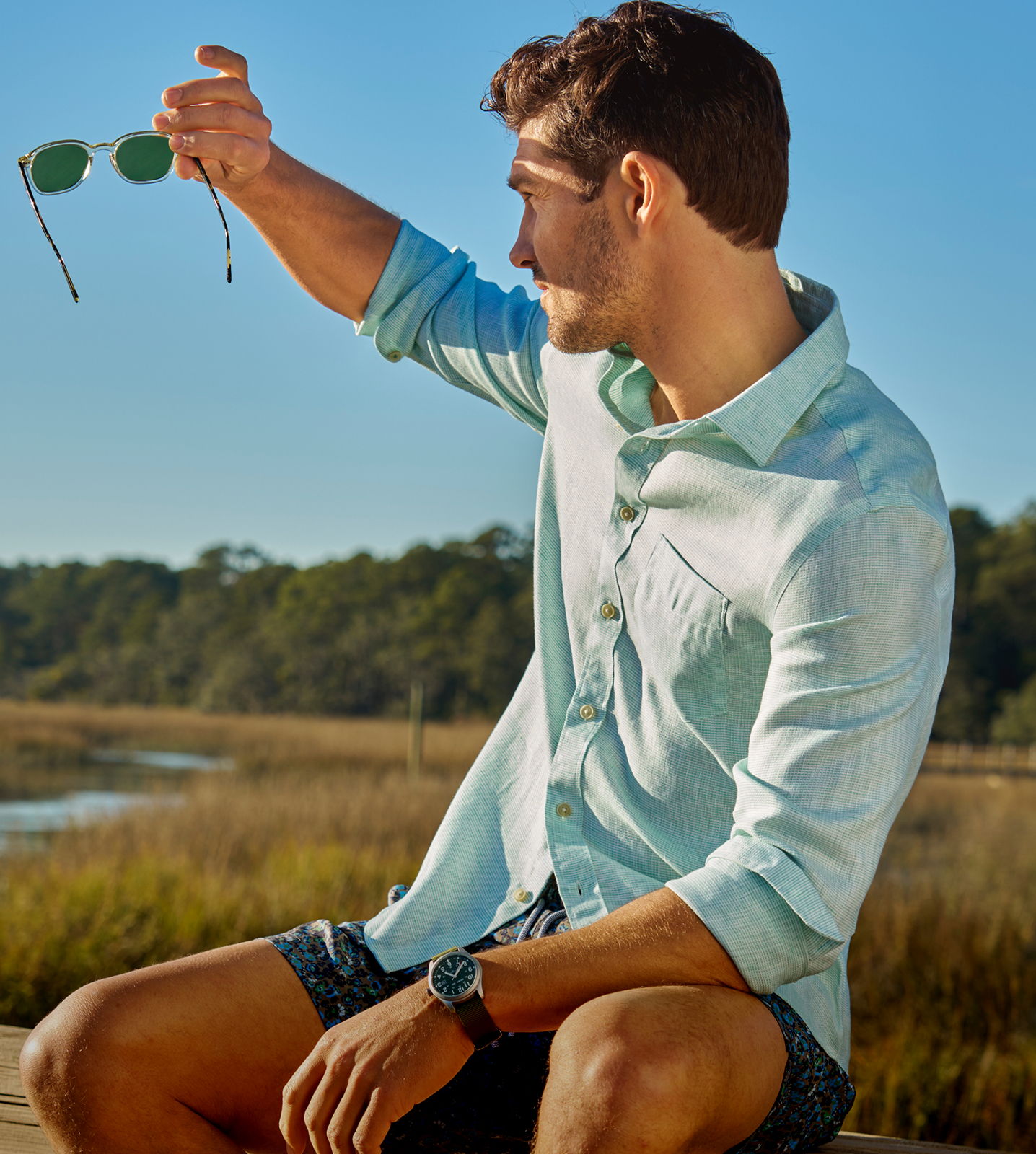 Young man holding sunglasses, sitting outdoors by a water body.