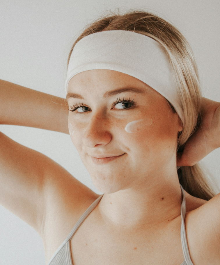 A smiling woman, hands behind her head, with a smear of facial cream on her left cheek